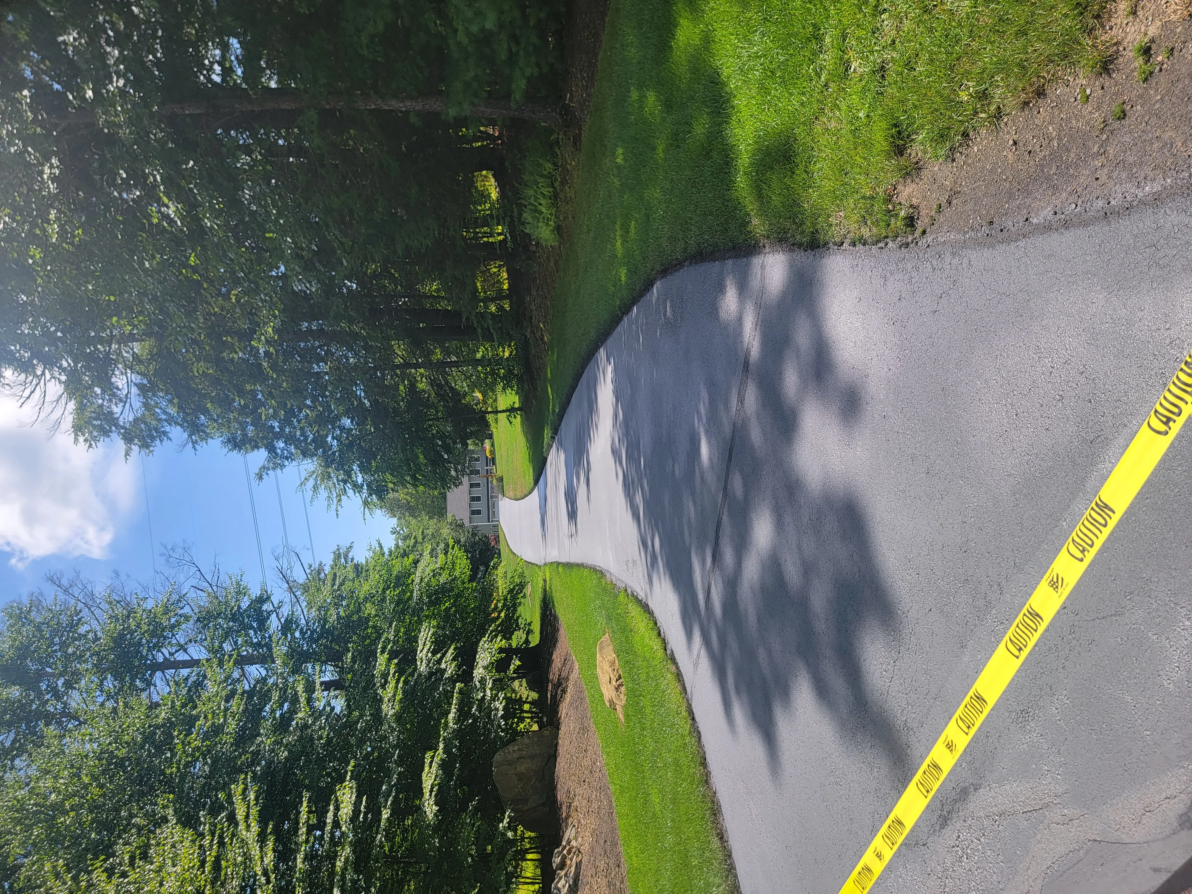 Freshly sealcoated asphalt driveway with dark finish, bordered by green lawn and mature trees
